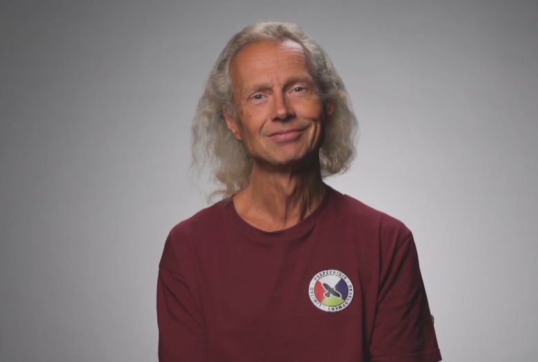 A man stands in front of a grey and white background. He is wearing a red shirt. 