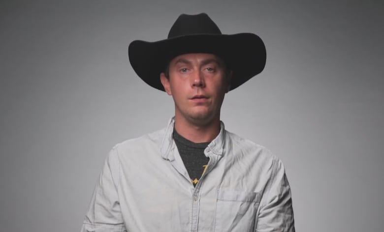 A man stands in front of a grey and white background. He is wearing a white button-up shirt and a cowboy hat.