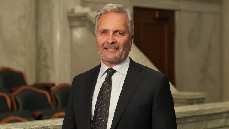 A man in a black suit and tie smiles to the camera.