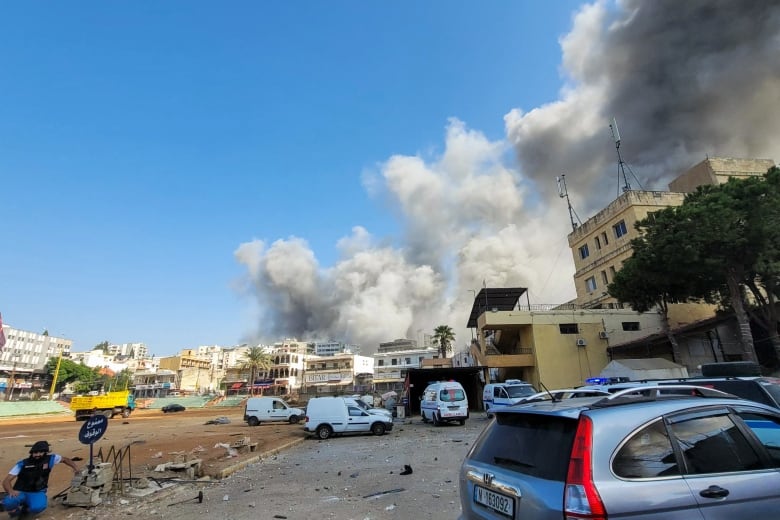 Several cars and multistorey low rise buildings are shown in the foreground as large clouds of grey smoke fill the air in the background.