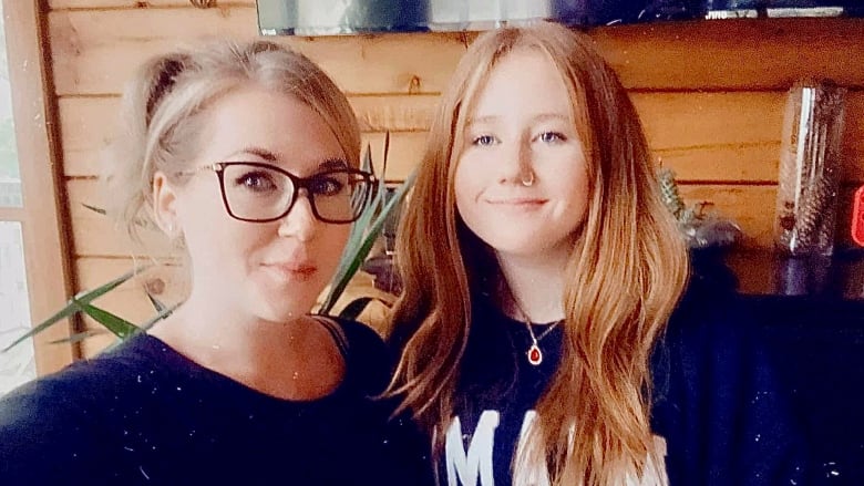 A woman with blonde hair pulled back in a ponytail smiles at the camera, while posed next to a younger girl in her early teens, with red hair, who is also smiling at the camera. 