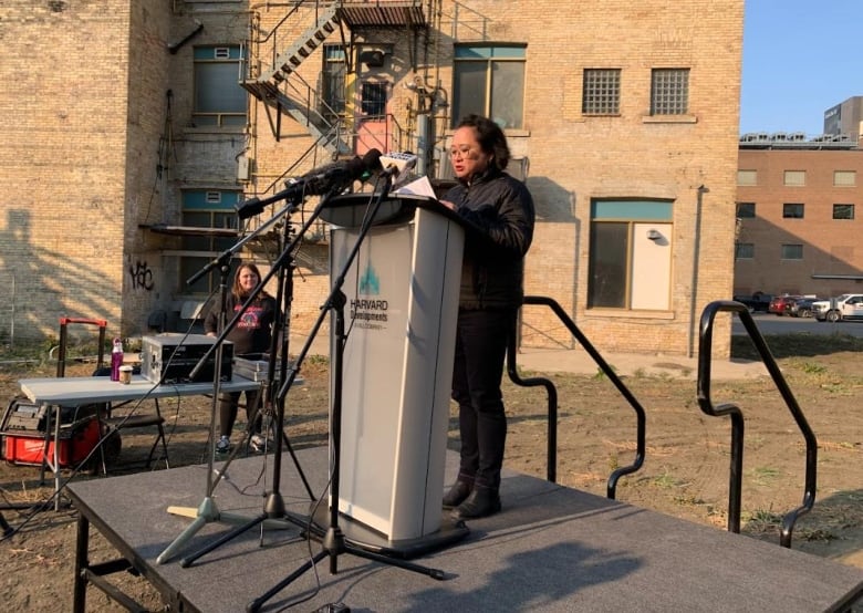 A woman is standing on a platform and reading on a stage from a microphone in the daylight. 