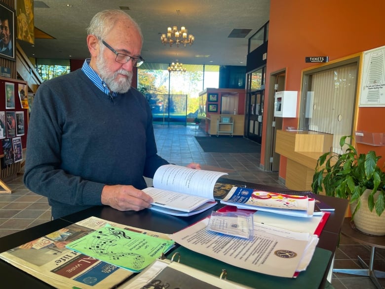 A bearded man wearing a blue sweater thumbs through a booklet placed on a high table.