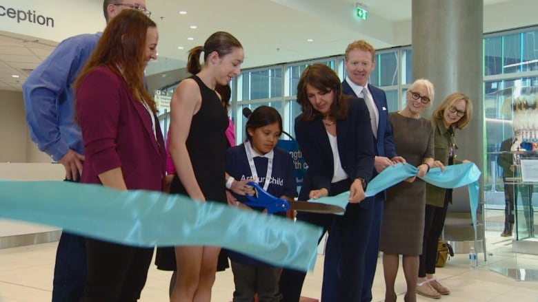 A group of people stand in a line, some of them hold a light blue ribbon while a young girl cuts it with large scissors.