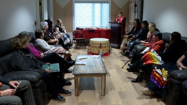 A group of Indigenous women sit in a common area. There is a large drum on the floor, and a woman in a red vest is speaking behind a podium. 