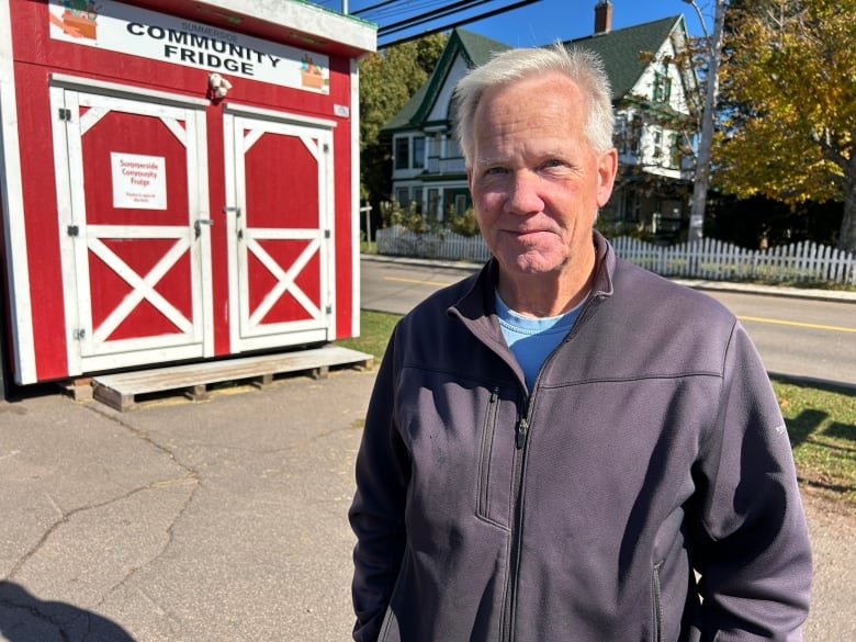 Man in blue jacket stands in front of a small red and white building with a sign saying Community Fridge. 