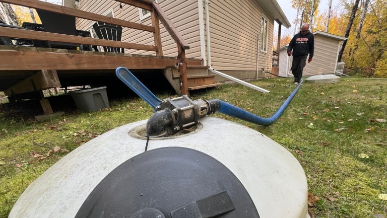 A man walks toward a water tank set in the ground, a large hose connected to fill it.