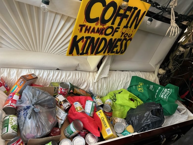 A real coffin filed with non-perishable food items such as canned food and Cheerios. A hand lettered sign reads 