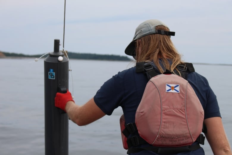 A female researcher has her back to the camera, she is on a boat facing the water. In her hand she holds a long black tube out over the water.