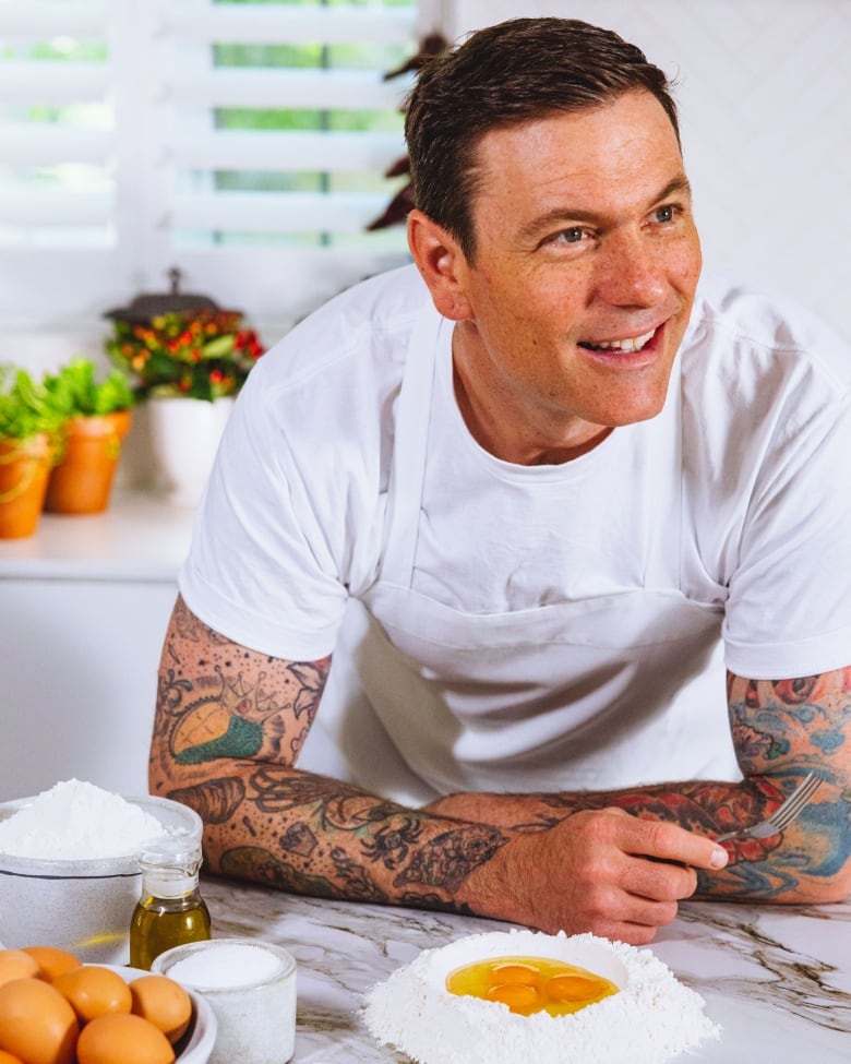 Promotional photo of an adult caucasian man leaning over a marble kitchen countertop. He's wearing a white tshirt and a white apron, and has tattoos on his arms.