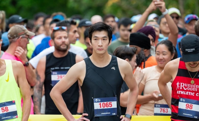 A man of East Asian descent in a racing bib, tank top, and shorts stands arms akimbo in a crowd of runners at the start of an outdoor race