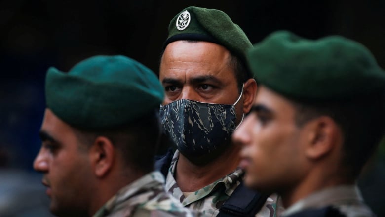 A masked soldier stands at attention. Two other soldiers, in the blurry foreground, march past.  