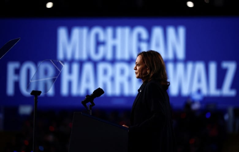 A woman with shoulder-length black hair wears a suit behind a podium. A large banner behind her reads 