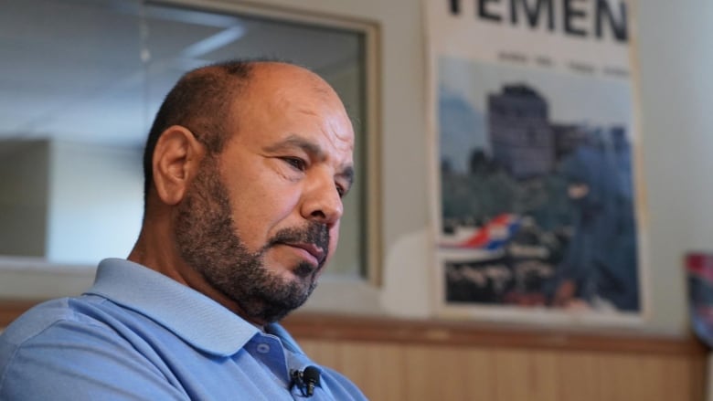 A man with black hair and black beard is seen wearing a blue polo shirt. A poster of Yemen is on the wall behind him.