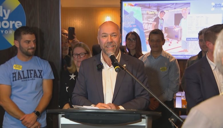 A smiling man wearing a suit standing behind a mic with a group of people crowded behind him. 