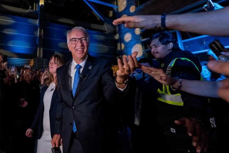 A man with white hair wears a dark suit and blue tie as he greets supporters.