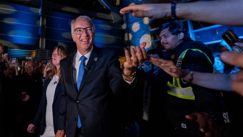 A man with white hair wears a dark suit and blue tie as he greets supporters.