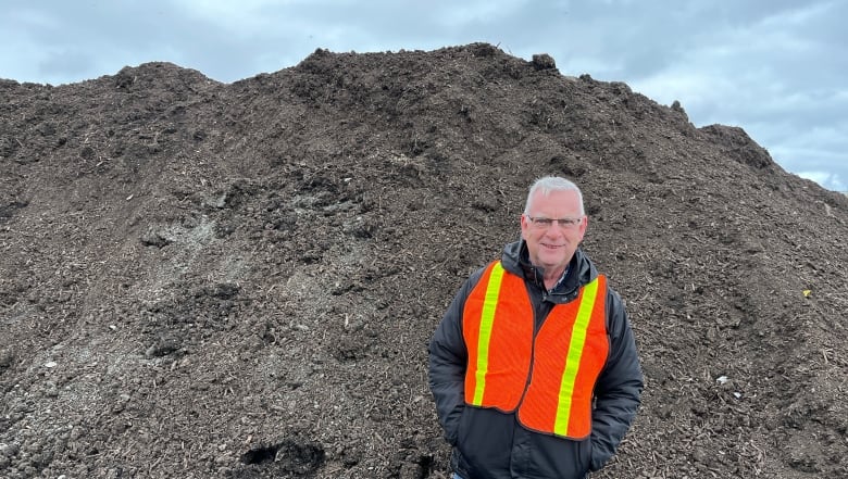 Man with short hair in a black jacket with a yellow and orange vest standing next to a mount of compost dirt.