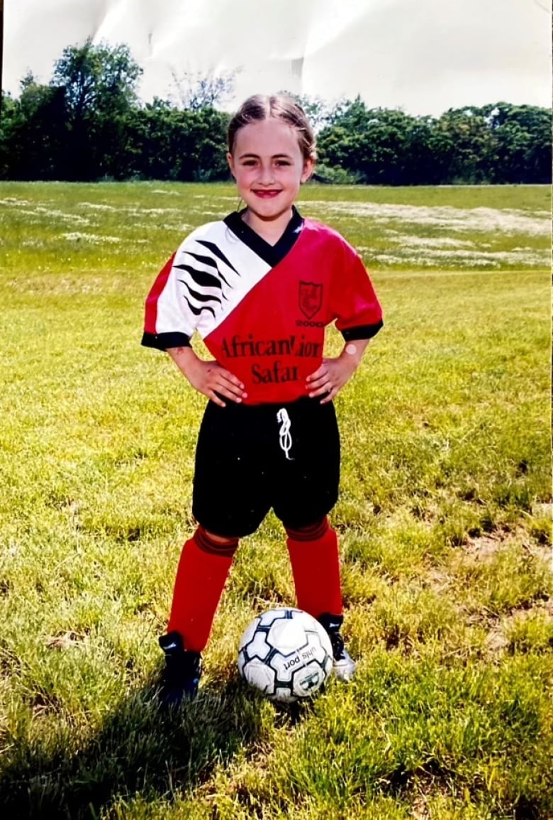 A young girl, wearong a full soccer kit, poses for a photo with a soccer ball at her feet.