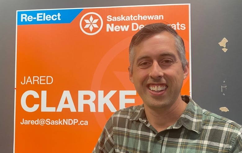 A grey-haired man in a plaid shirt stands before a bright orange sign that reads 'Jared Clarke.' 