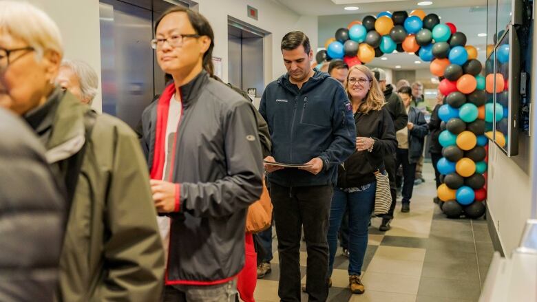 several people stand in a queue with balloons in an arch in a hallway