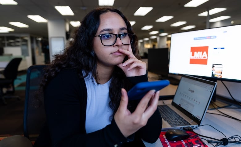 A young woman holds a blue cell phone, while a screen in the background shows an advertisement for an LMIA.