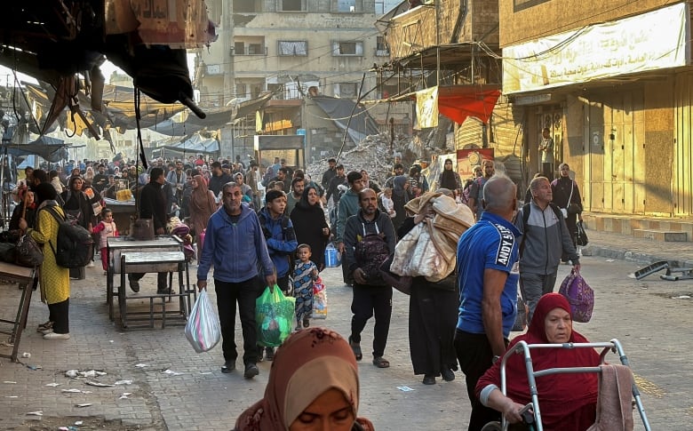 A crowd of people, young and old, walk on a street carrying bags of belongings.