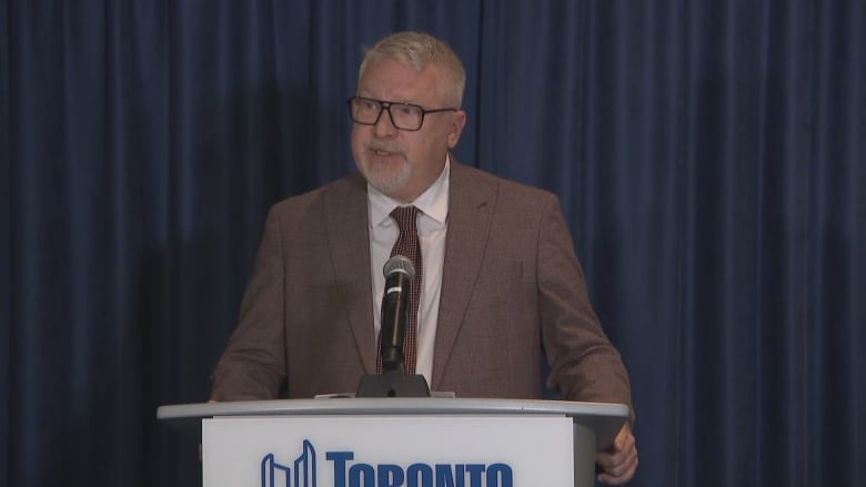 A middle aged white man in a suit and tie speaks at a City of Toronto Podium into a microphone. The background is a blue curtain