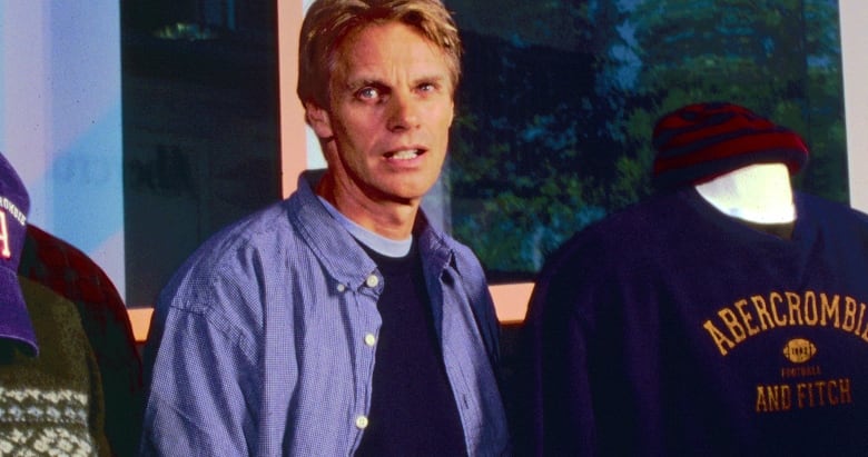 An older photo shows a man in a blue un-buttoned button-up, standing in between two mannequins, one of which is wearing a sweater that says Abercrombie & Fitch on it. 