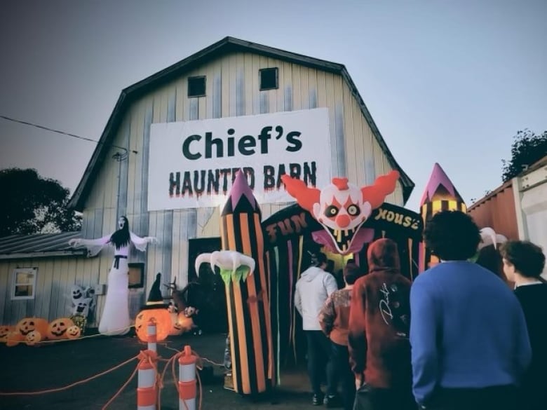 Sign on old barn says 'Chief's Haunted Barn' with pumpkins and ghosts in front on barn, scary clown blow-up entrance with people filing in. 