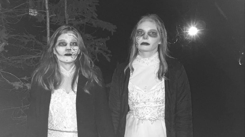 Two women with medium-length hair dressed in long white lacy dresses and dark coats, wwith scary white makeup on their faces. 