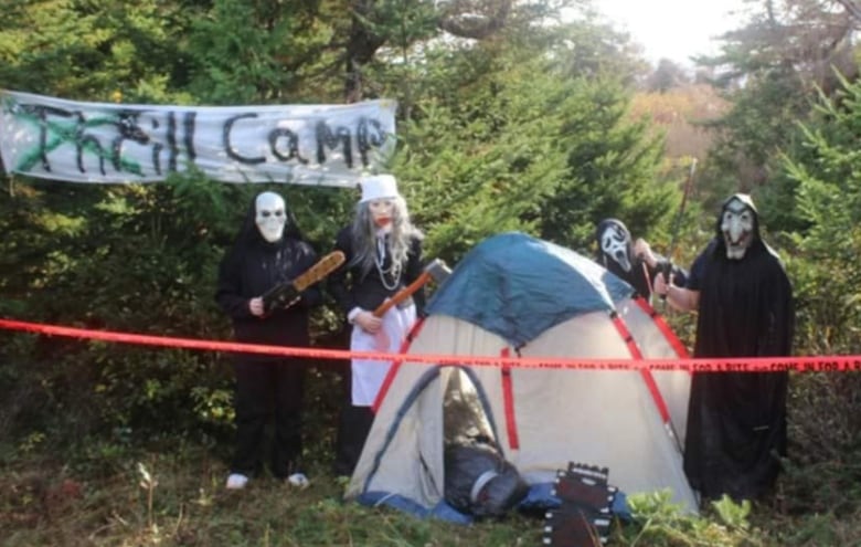 Volunteer actors dressed in scary masks and costumes holding a chain saw and axes, surround a tent in the woods with a sign that says 