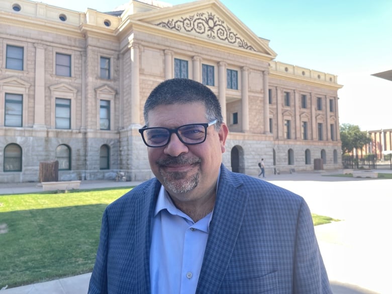 A man wearing glasses and a goatee is pictured in front of a government building.