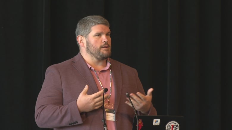 Image of man in a burgundy blazer speaking at a microphone.