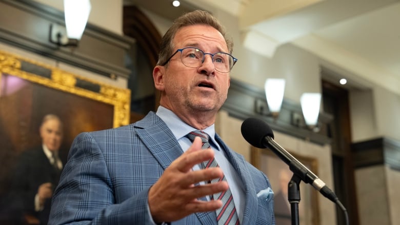 A stately man speaks at a microphone with the portrait of a Canadian Prime Minister in the background.