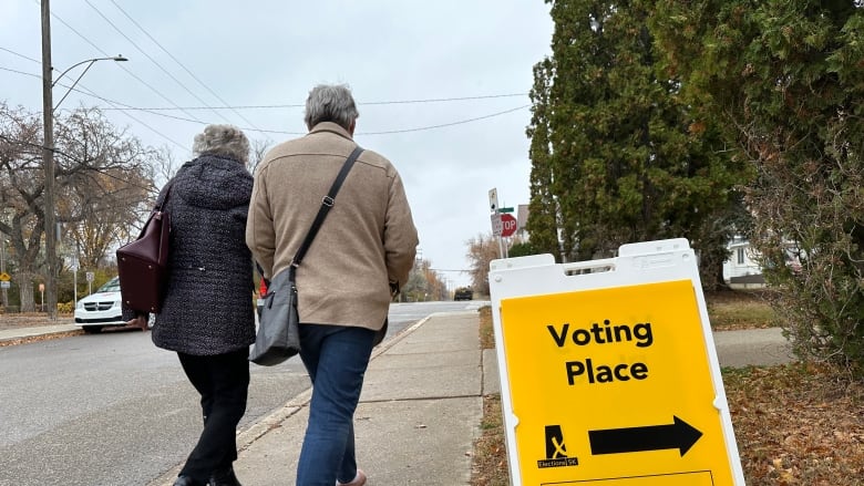 Two people walk away from the camera, down a sidewalk. A yellow sign sits. It has the phrase 