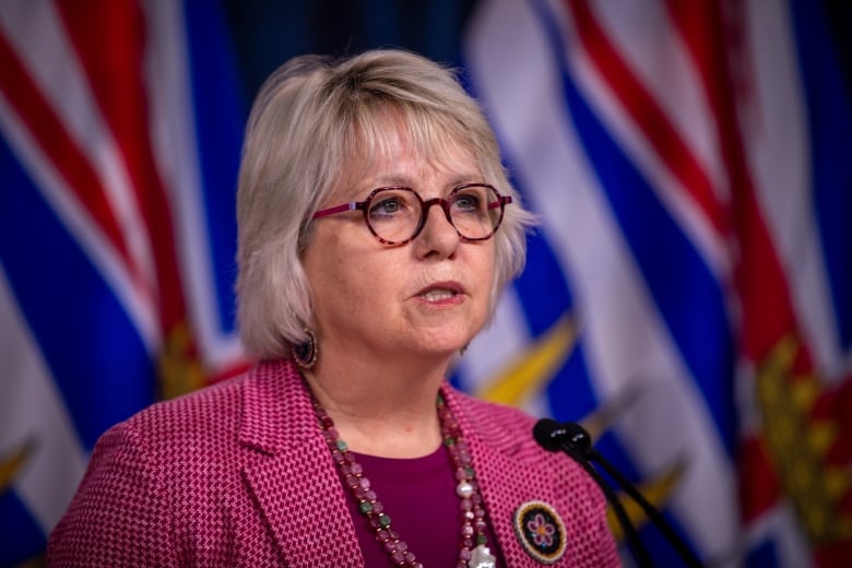 A woman wearing a pink sweater speaks in front of B.C. flags.