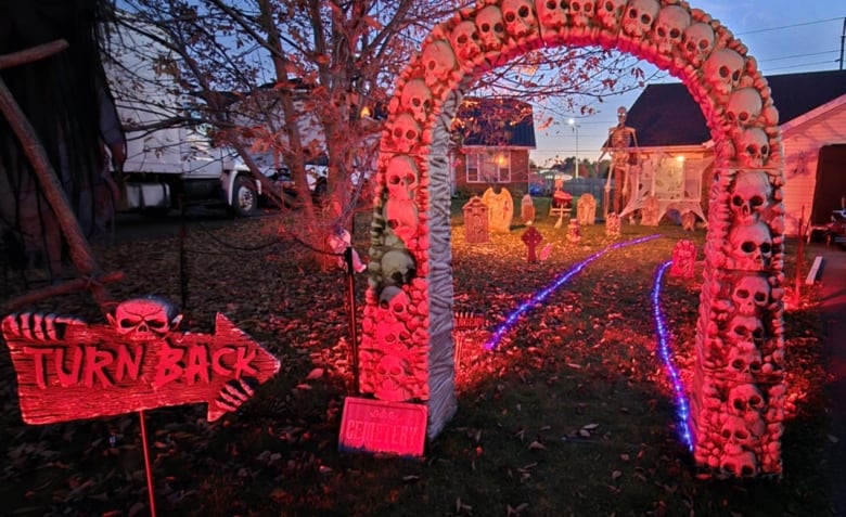 Exterior of house decorated for Halloween, featuring an arch of skulls, a sign that says Turn Back, fake gravestones and skeleton in background, lit with red lights. 