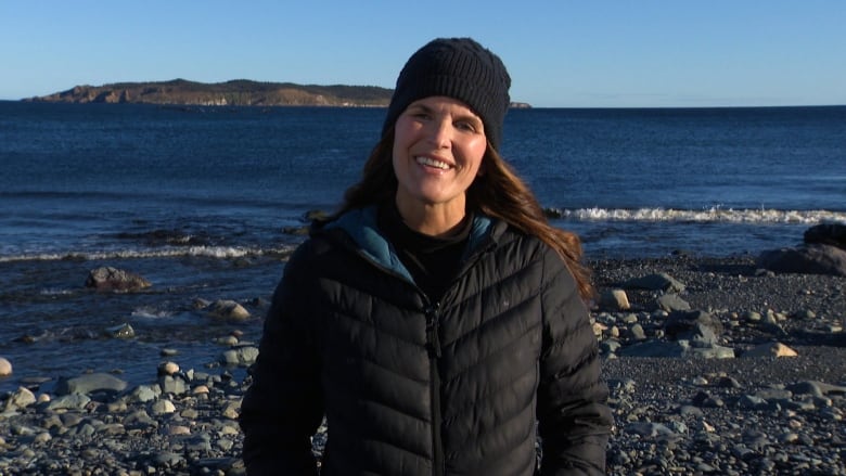 A woman wearing a puffer jacket and a toque stands on a rocky beach in front of the Atlantic Ocean.