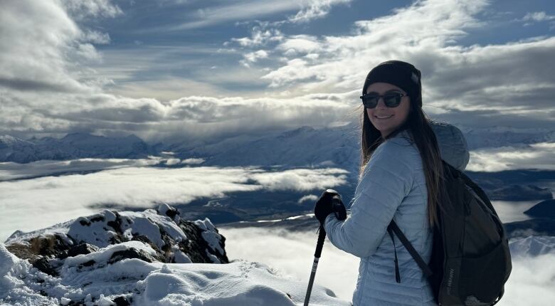 A woman on a snowy mountain wearing in glasses and a beanie smiles for a photo.