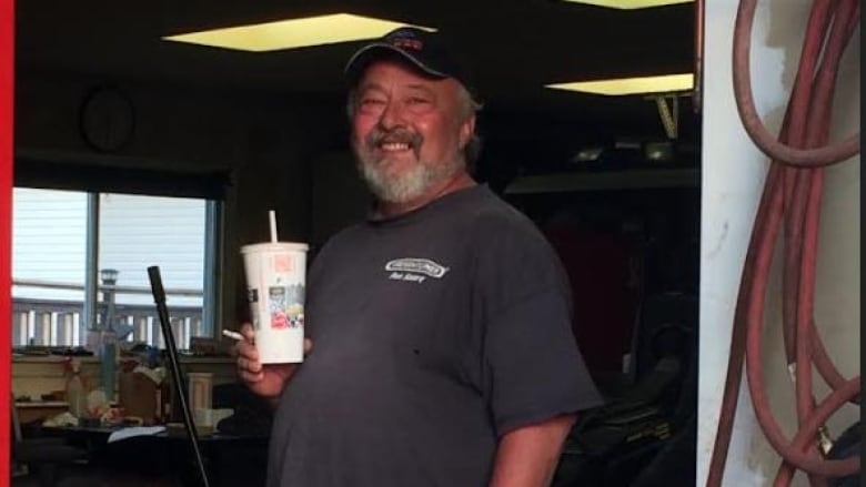 An older gentleman standing in a garage holding a drink with a smile on his face.