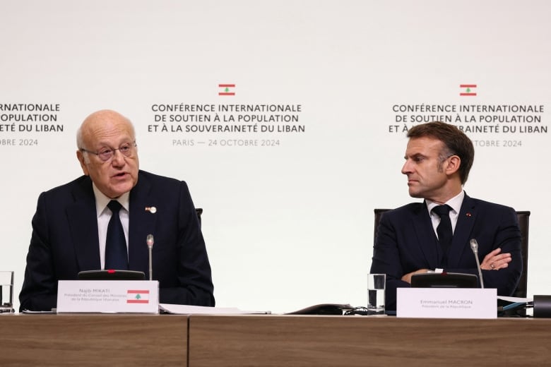 A balding, bespectacled, cleanshaven man in a suit and tie sits at a panel table and speaks while a younger cleanshaven man in suit and tie listens with arms folded.