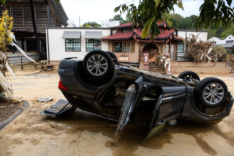 Overturned car on dirt covered road with buildings in background