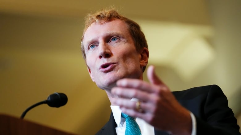 A man in a black suit and turquoise tie gestures with his hand as he speaks into a microphone.