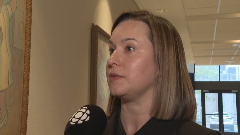 A woman with short brown hair stands in front of a CBC microphone.