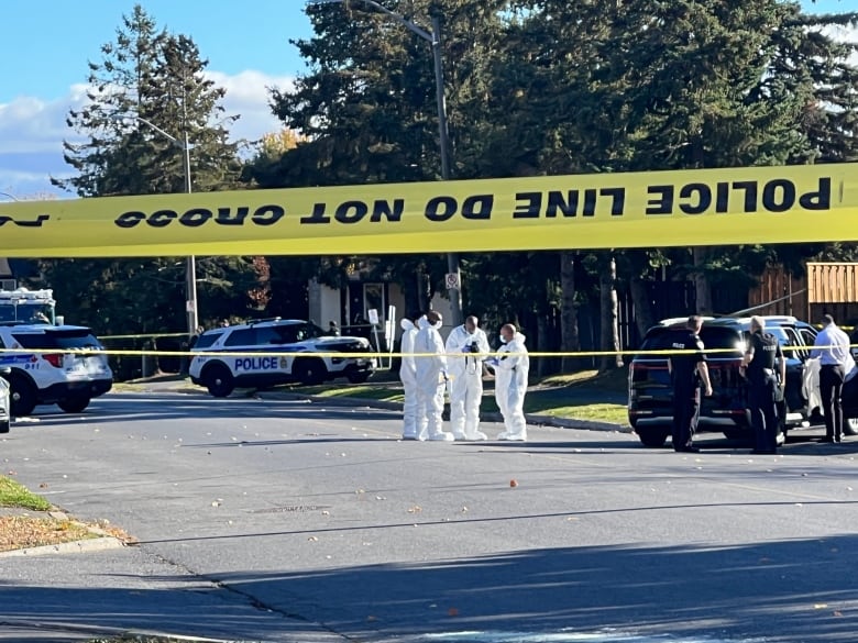 A suburban park taped off with police tape. People stand around in white suits.