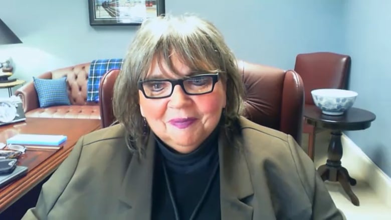 A white woman with shoulder-length grey hair and black glasses wears an olive blazer and black shirt. She sits at a desk with an office behind her