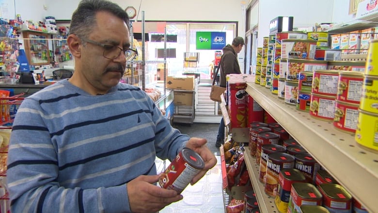 A shop owner looks at a can of food while standing in an aisle of his store. 