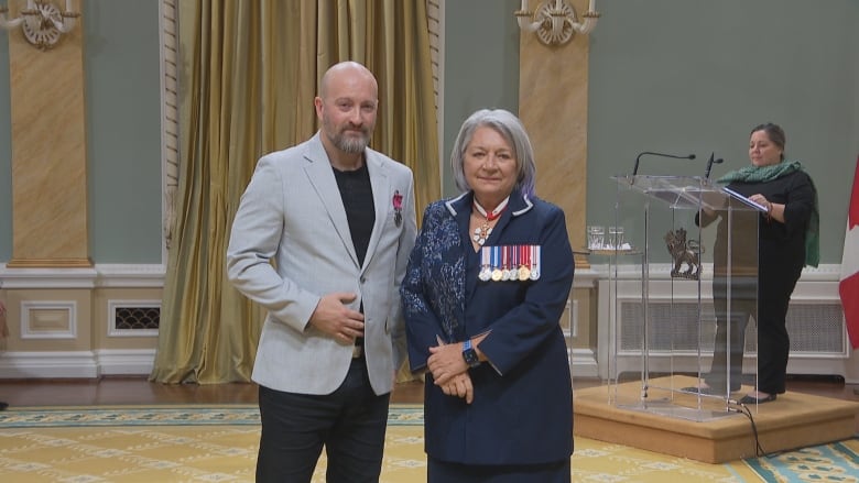 A man wearing a grey blazer and a medal over his heart stands with a woman wearing a navy suit and medals.