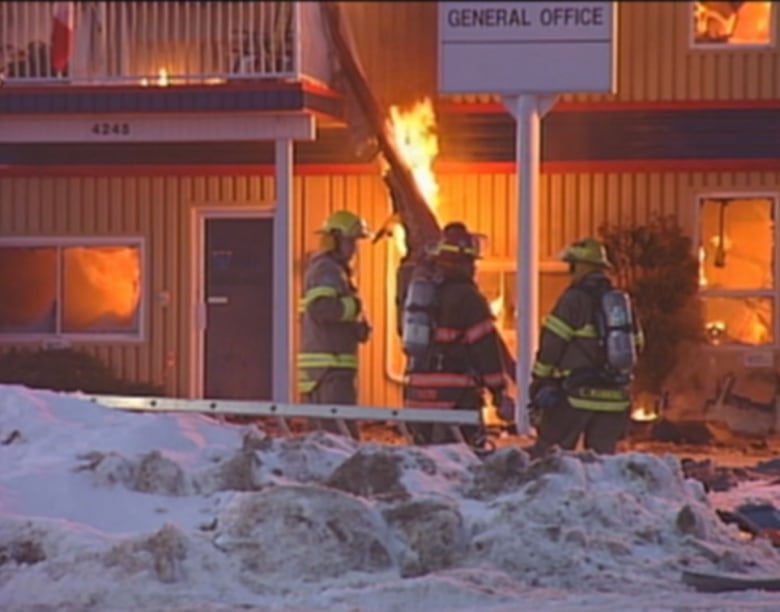 Three firefighters stand around a burning piece of debris at a building with snow piled up